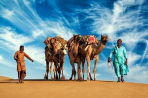 Two camels and two men in the desert with an incredible sky above them