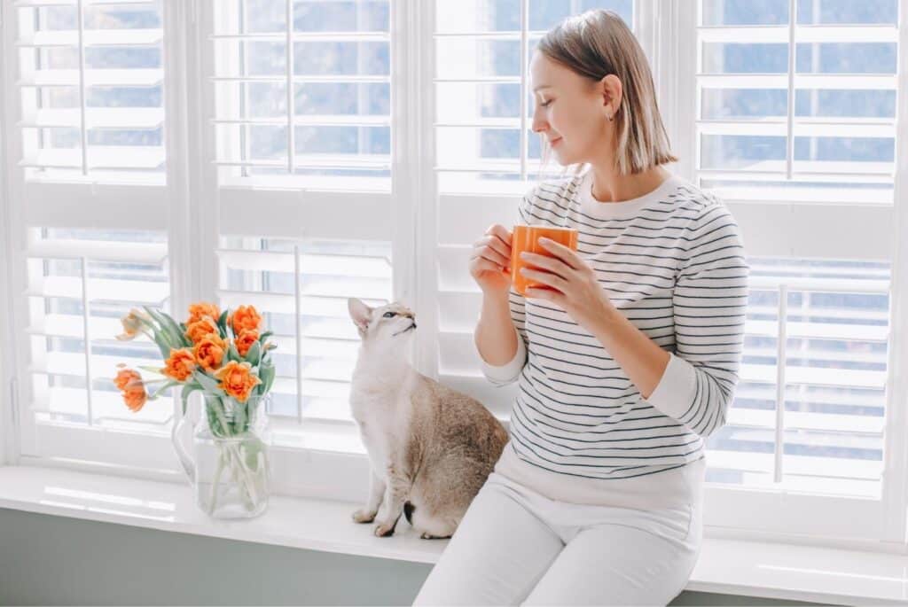 Woman sitting by her window drinking key with her cat - annakraynova - canva pro